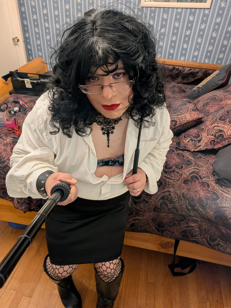 Mistress Ilya is standing by a bed with a riding crop in her hand.