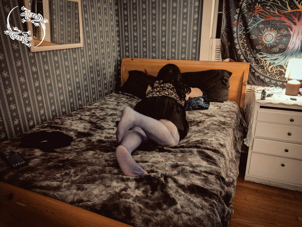 Mistress Ilya is laying on her bed with her back to the camera. 