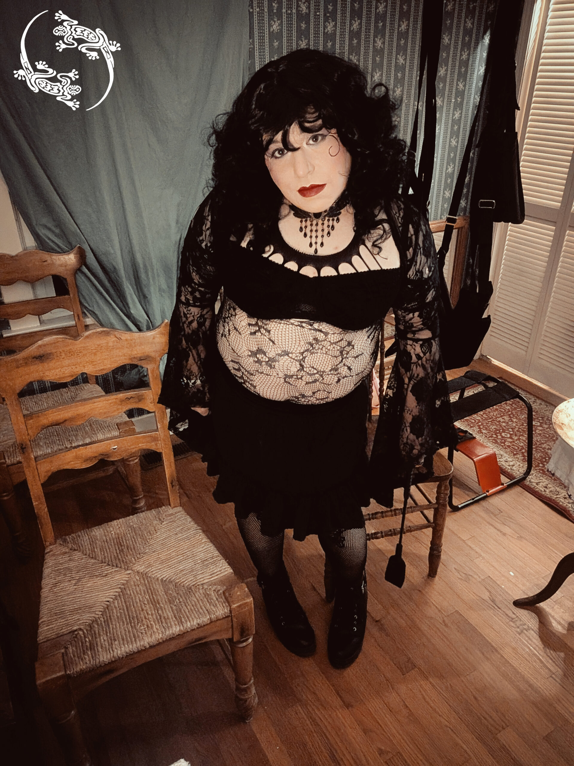 Mistress Ilya is standing by some chairs with a riding crop in her hands. 