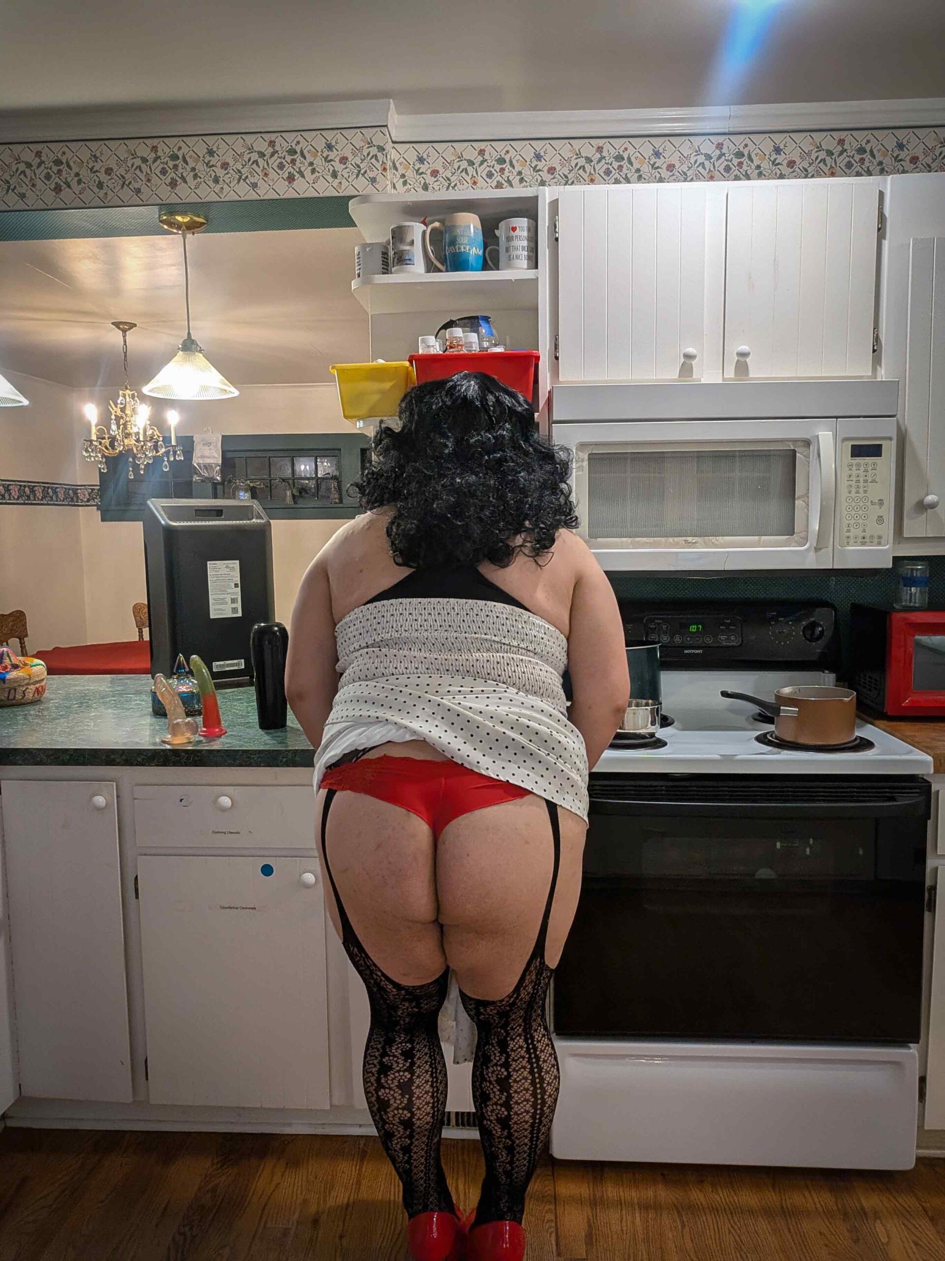 Mistress Ilya is in her kitchen with her ass showing under her dress. She is leaning against a stove with her back to the camera. 