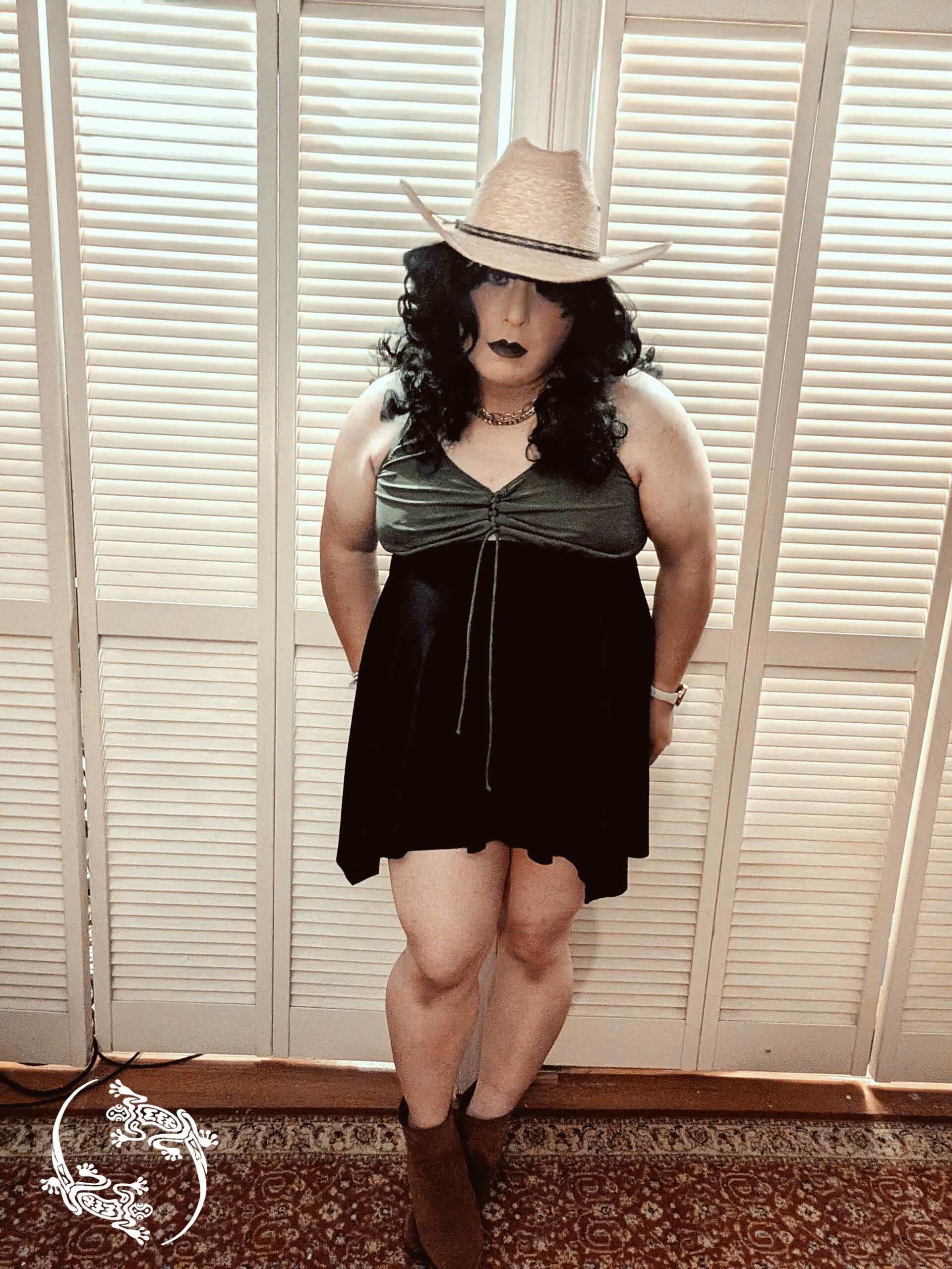 Mistress Ilya is wearing boots, a cowboy hat, and a swimsuit. She is standing up.