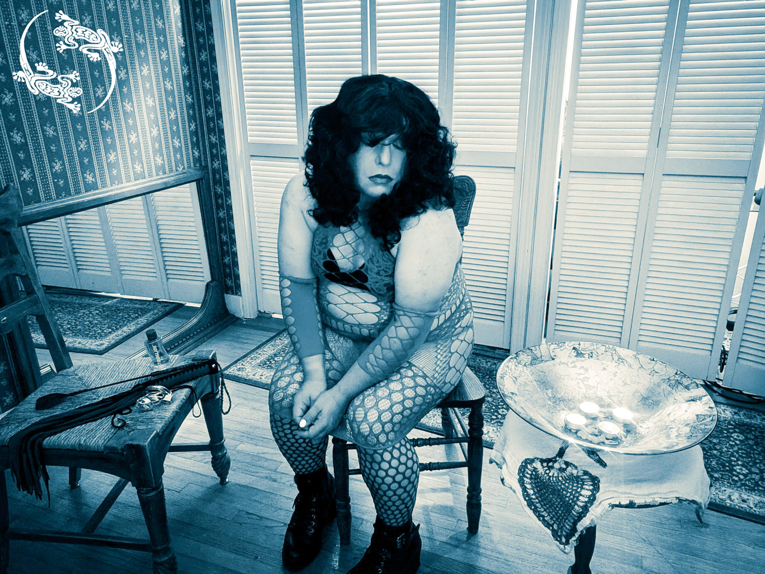 Mistress Ilya is sitting on her chair with her hands between her legs. Candles burn on a plate next to her.