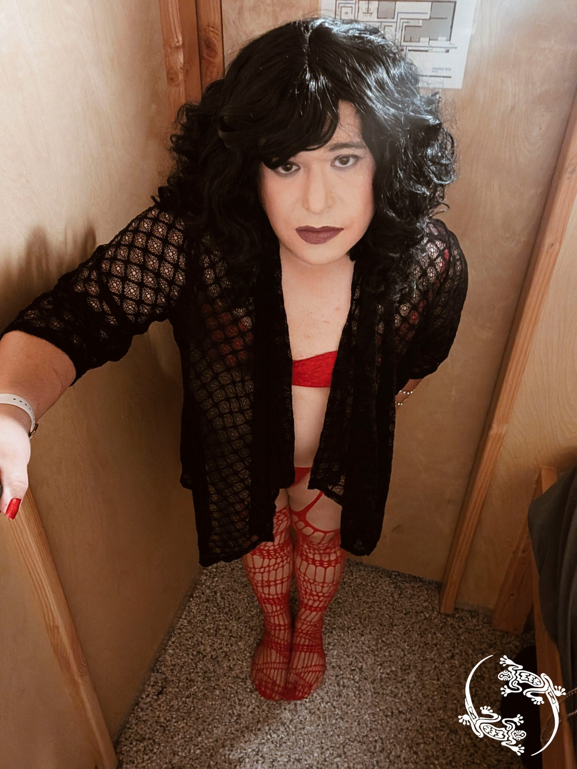 Mistress Ilya is standing in a small room wearing a lacy top and lingerie. 