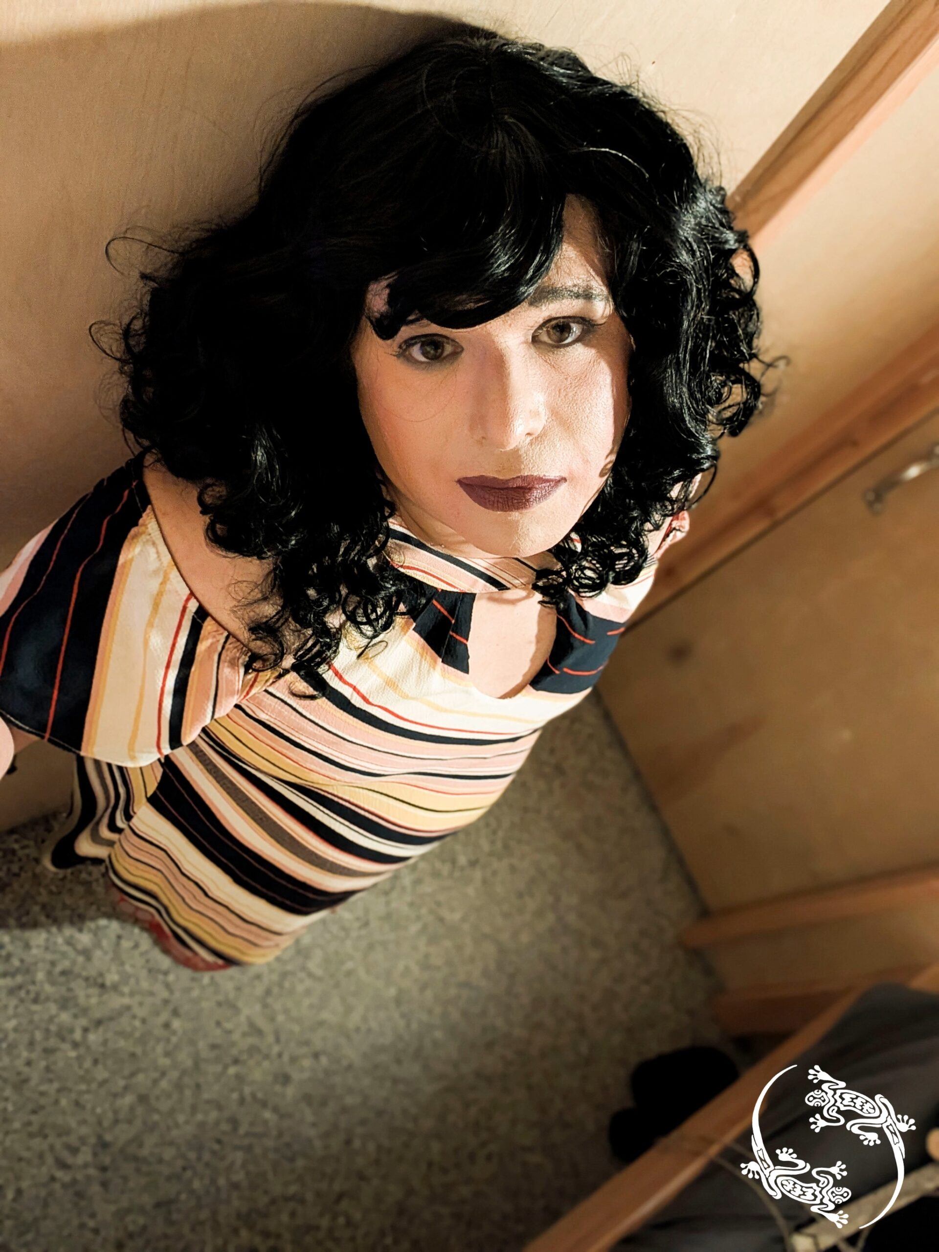 Mistress Ilya is standing with her back against a wall in a small room.