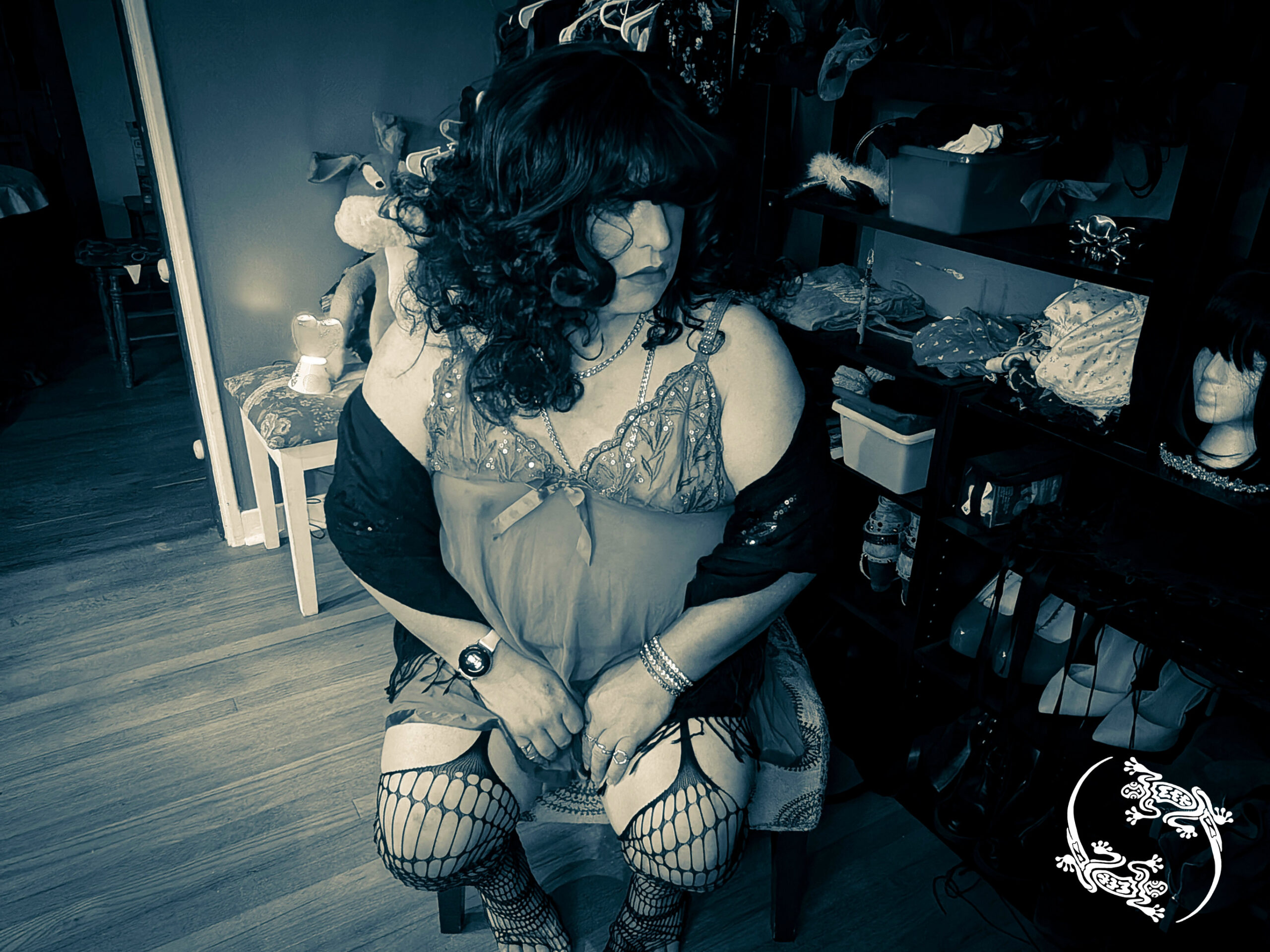 Mistress Ilya is sitting on a chair next to a shelf with clothing accessories. 
