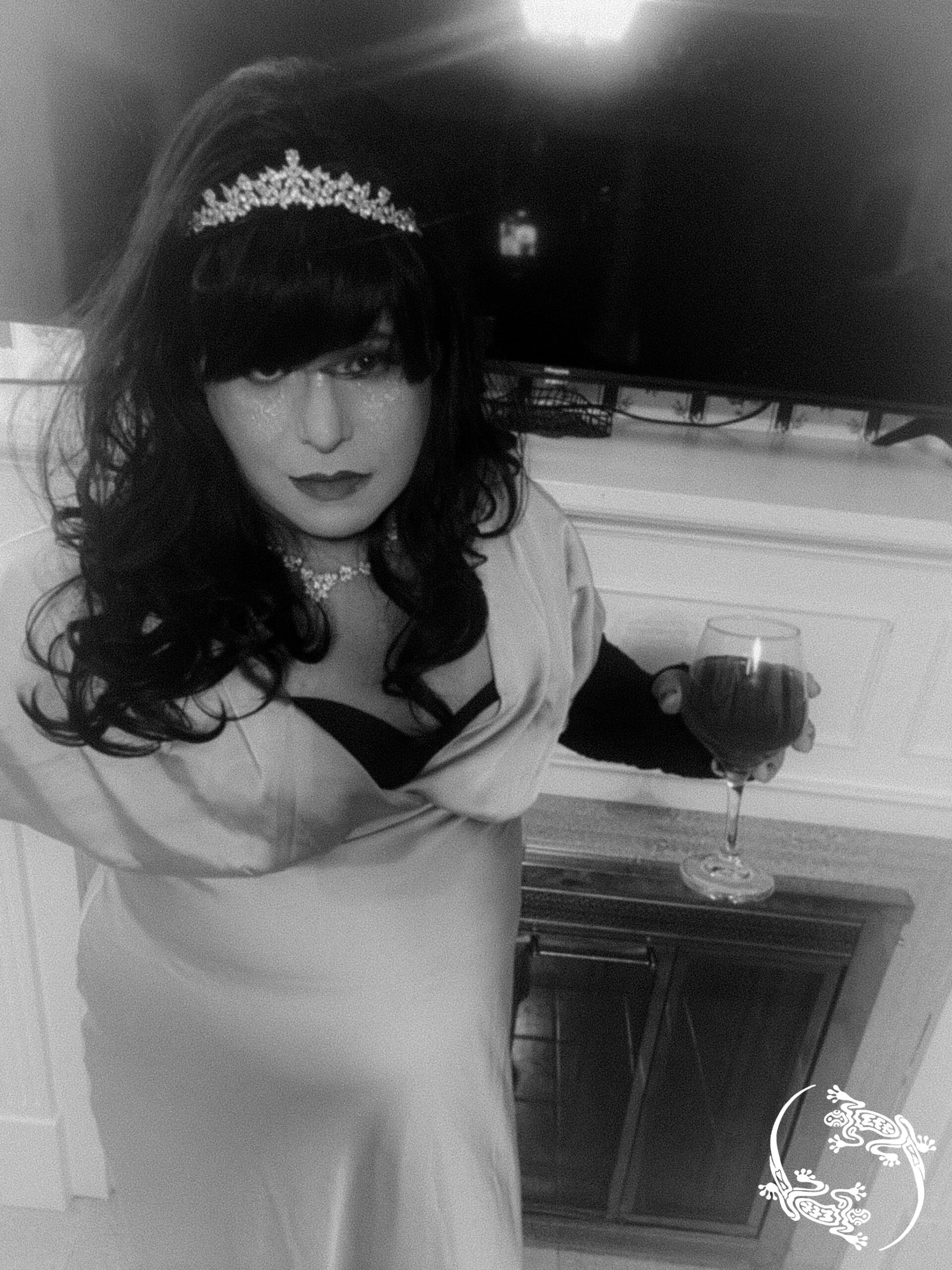 Mistress Ilya is wearing a crown and a dress while holding a wine glass. She is standing next to a fireplace. 
